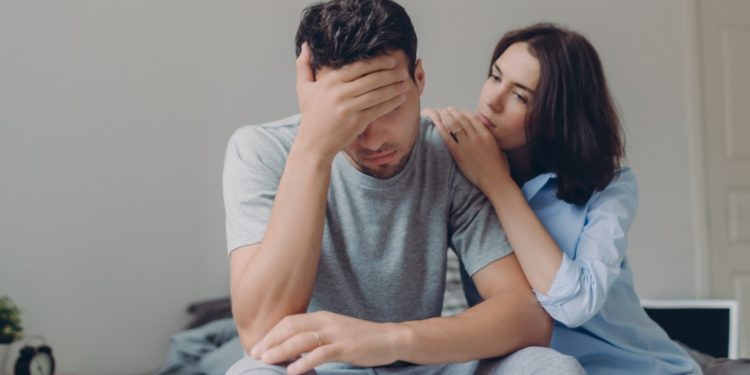 tadalafila - homem sentado na cama com a mão na cabeça, mulher consolando apoiada no ombro dele